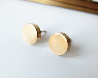 May include: Two gold-colored, round stud earrings. Each earring has a flat, circular face and a post for insertion. The earrings are set against a white background, with a gold-colored frame visible in the background.