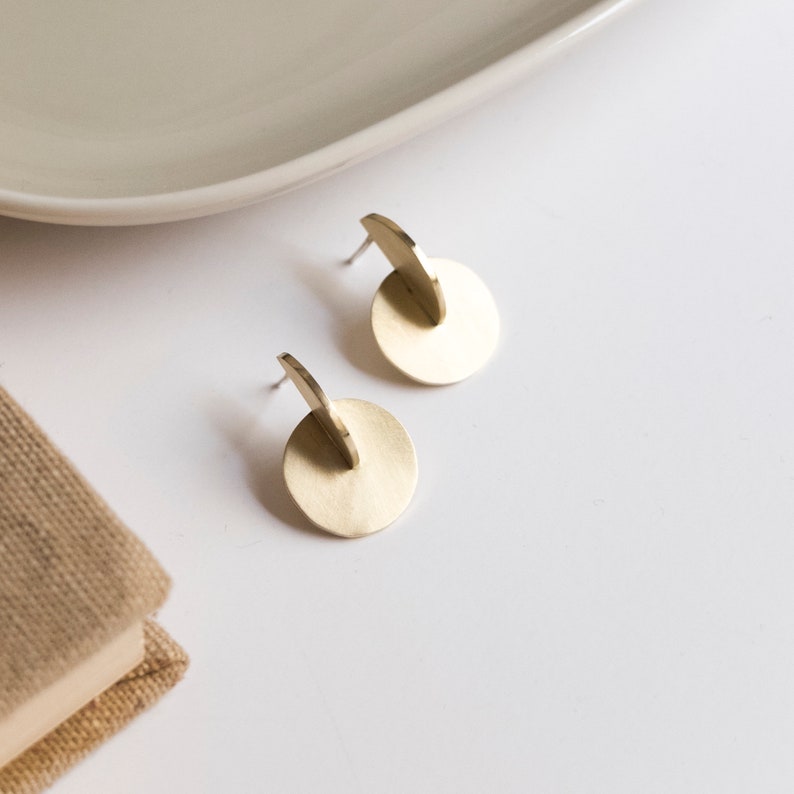 May include: A pair of gold-toned stud earrings featuring a flat circular disc design with a perpendicular semi-circular metal piece attached to the center. The earrings have a brushed matte finish and include standard metal earring posts on the back.