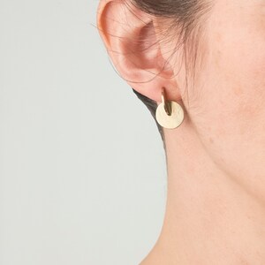 May include: A gold-toned earring featuring a small, smooth circular disc suspended from a curved, hook-like metal piece. The earring has a polished, reflective surface and a simple, geometric design that hangs directly from the earlobe.