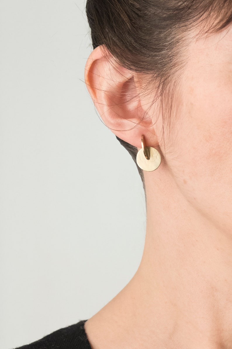 May include: A gold-toned earring featuring a small, smooth circular disc suspended from a curved, hook-like metal piece. The earring has a polished, reflective surface and a simple, geometric design that hangs directly from the earlobe.