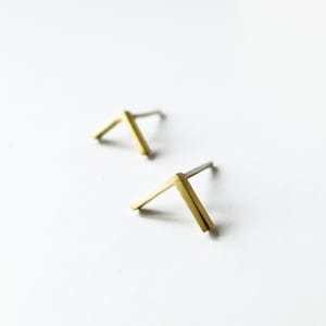 May include: Two minimalist gold-colored stud earrings in an angled V-shape, with silver posts. The earrings are set against a clean white background, creating a modern and simple aesthetic. The earrings are small and delicate.