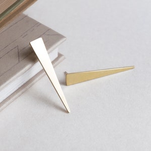 Puede incluir: Dos horquillas triangulares de color dorado. Una horquilla está de pie, mientras que la otra está horizontal. Las horquillas están sobre una superficie clara, con una pila de libros en el fondo.