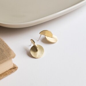 May include: A pair of gold-toned stud earrings featuring a brushed metal finish. Each earring consists of a flat circular disc intersected by a smaller semi-circular piece positioned vertically, creating a three-dimensional geometric structure.