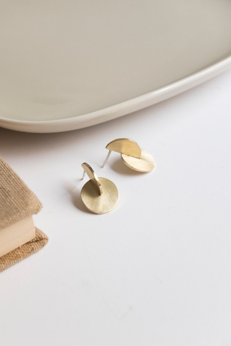 May include: A pair of gold-toned stud earrings featuring a brushed metal finish. Each earring consists of a flat circular disc intersected by a smaller semi-circular piece positioned vertically, creating a three-dimensional geometric structure.