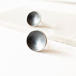 Könnte beinhalten: Zwei silberfarbene Ohrstecker, jeweils in Form einer Halbkugel. Die Ohrringe haben ein konkaves Design mit dunklem Innenraum und hellem, reflektierendem Außenrand. Sie sind vor einem weißen Hintergrund platziert, was eine minimalistische Ästhetik erzeugt.