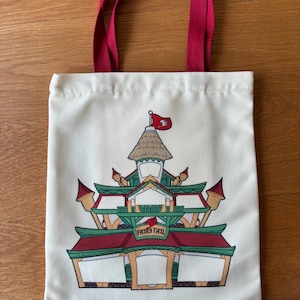 May include: Off-white tote bag with red handles. The bag features a colorful illustration of a building with a red roof, green accents, and the text "FAIRY TAIL". A red flag waves from the top of the building.