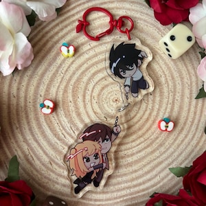 Op de afbeelding: Twee acryl sleutelhangers met anime-personages en een rode, appelvormige sluiting. Eén sleutelhanger toont een personage met zwart haar, de andere toont twee personages die elkaar omarmen. Decoratieve appelvormige kralen en een dobbelsteen zijn verspreid.