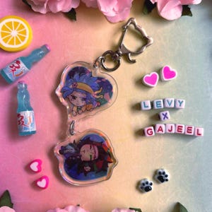 May include: A clear acrylic keychain featuring two anime-style character designs within heart-shaped frames. The keychain is attached to a silver clasp. Additional elements include miniature blue soda bottles, pink heart-shaped beads, and the text "LEVY x GAJEEL".