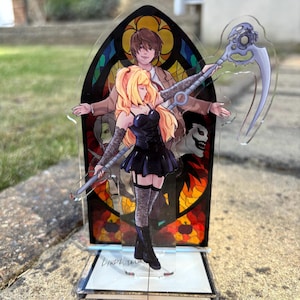 May include: A clear acrylic stand featuring anime-style artwork. The design includes a woman in a black dress holding a scythe, with a stained-glass window background. The base of the stand has the text "LINDA TAIL".