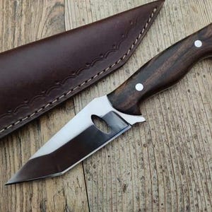 May include: A brown leather sheath with a decorative stitching pattern, holding a silver knife with a dark wood handle. The knife has a small hole in the blade and a rounded tip.