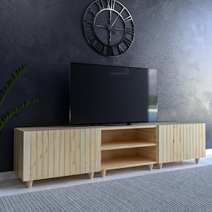 May include: A light-coloured wooden TV stand with vertical plank detailing, featuring two doors and an open shelf area. A large black flat-screen TV sits atop the stand. A decorative metal clock hangs on the dark wall behind the TV.
