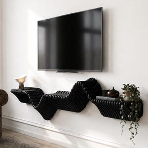 May include: A black wall-mounted floating shelf with a wavy, layered, and ribbed texture. The shelf is positioned directly beneath a flat-screen television mounted on a white wall, featuring flat surfaces on the ends for decorative items.