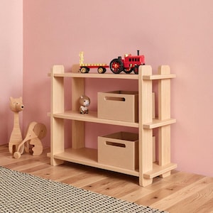 May include: A three-tiered, light-wood bookshelf with two beige storage boxes and toy vehicles. A child in a camouflage shirt and green trousers stands nearby. Wooden giraffe and dog ornaments are on the floor.