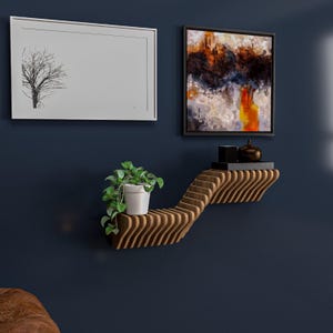 Pu&ograve; includere: Un ripiano a muro in legno con un design ondulato. Il ripiano &egrave; montato su una parete blu scuro e ha una piccola pianta in vaso e due oggetti decorativi.