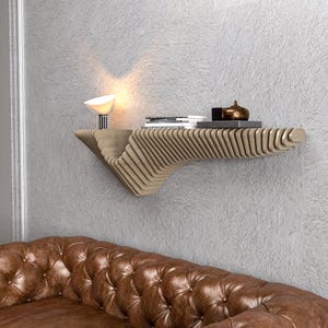 Pode incluir: Uma prateleira de parede de madeira clara com um design curvo. A prateleira est&aacute; montada em uma parede cinza e tem uma pequena l&acirc;mpada e alguns livros.