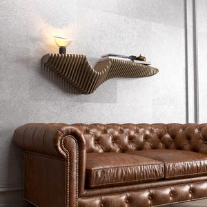 Pode incluir: Um sof&aacute; de couro marrom com capiton&ecirc; de bot&otilde;es fica em frente a uma parede com uma prateleira de madeira. A prateleira &eacute; curva e tem um design &uacute;nico. Uma l&acirc;mpada fica na prateleira.