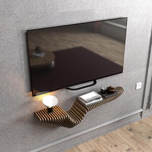 Pode incluir: Uma prateleira de parede de madeira moderna com um design ondulado. A prateleira &eacute; montada em uma parede cinza e tem uma televis&atilde;o preta plana montada acima. H&aacute; uma pequena l&acirc;mpada e alguns livros na prateleira.