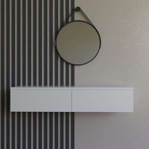 May include: A contemporary wall-mounted unit with a circular mirror. The white vanity has two drawers, and the mirror is framed in black. The background includes a vertical striped feature wall in grey and a neutral-toned wall.