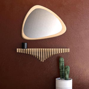 May include: A modern wall decor arrangement featuring a triangular mirror with a light wood frame, a slatted wooden shelf, and a potted cactus. The mirror is above the shelf, and the cactus is in a white pot. The wall is a deep reddish-brown.