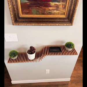 Pu&ograve; includere: Un ripiano a muro in legno con un design ondulato. Il ripiano &egrave; in legno marrone chiaro e ha una finitura naturale. Ci sono tre piccole piante sul ripiano, due in vasi bianchi e una in un vaso marrone. C'&egrave; un piccolo vassoio nero sul ripiano con il testo "Be the Change" in lettere dorate.