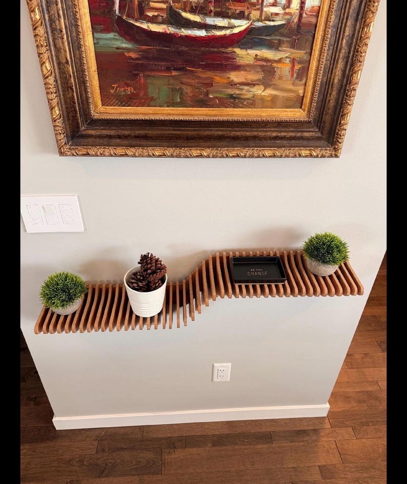 Pu&ograve; includere: Un ripiano a muro in legno con un design ondulato. Il ripiano &egrave; in legno marrone chiaro e ha una finitura naturale. Ci sono tre piccole piante sul ripiano, due in vasi bianchi e una in un vaso marrone. C'&egrave; un piccolo vassoio nero sul ripiano con il testo "Be the Change" in lettere dorate.