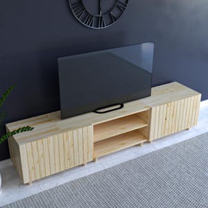 May include: A light-colored wooden TV stand with a modern design. The stand has two side cabinets and an open center section with two shelves. A large black TV sits on top. The stand is approximately 72 inches long.