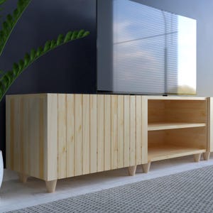 May include: A light-colored wooden TV stand with a vertical slat design on the front. The stand has a rectangular shape with short, angled legs. It features an open shelf area with two shelves and is positioned beneath a large television.