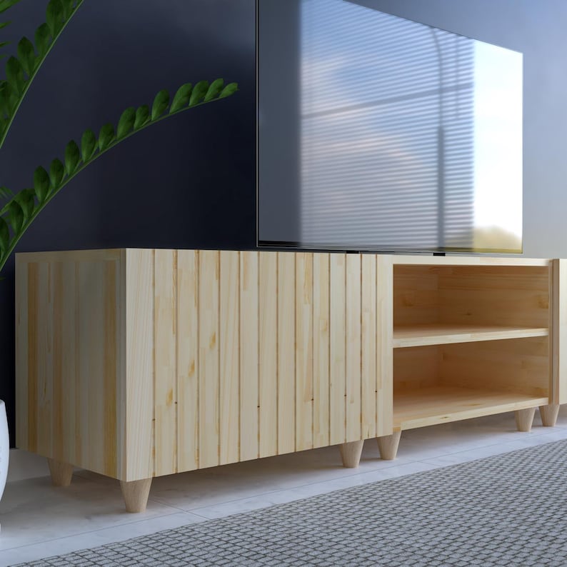 May include: A light-colored wooden TV stand with a vertical slat design on the front. The stand has a rectangular shape with short, angled legs. It features an open shelf area with two shelves and is positioned beneath a large television.