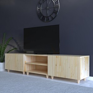 May include: A light-colored wooden TV stand with a modern design. The stand has two doors and an open shelf area. The stand is supported by short, rounded legs. A large black television sits on top of the stand.