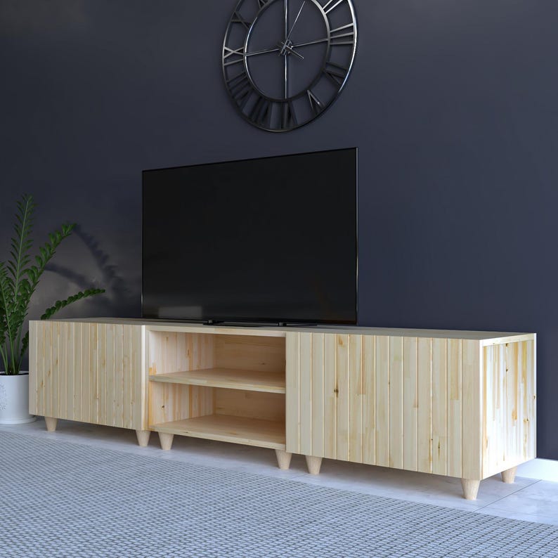 May include: A light-colored wooden TV stand with a modern design. The stand has two doors and an open shelf area. The stand is supported by short, rounded legs. A large black television sits on top of the stand.