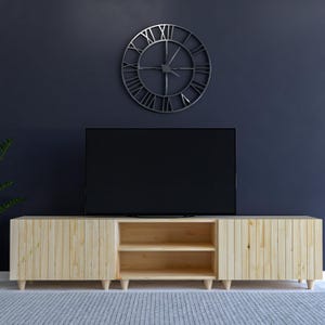 May include: A light-colored wooden TV stand with two doors and an open shelf section. The stand is set against a dark blue wall, with a large metal clock and a black television above it. The stand has a mid-century modern design.