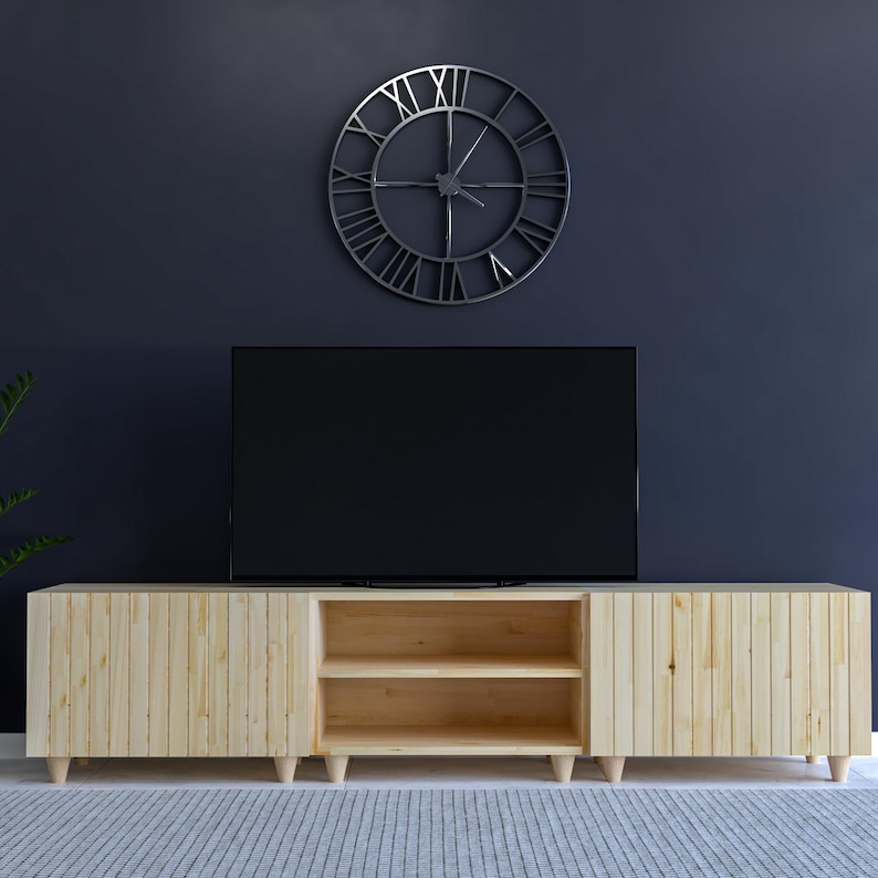May include: A light-colored wooden TV stand with two doors and an open shelf section. The stand is set against a dark blue wall, with a large metal clock and a black television above it. The stand has a mid-century modern design.