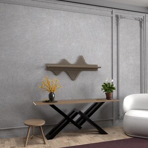 Op de afbeelding: Een moderne consoletafel met een zwart X-vormig onderstel en een lichtbruin houten blad. De tafel is versierd met een vaas met gele bloemen en een potplant. Een witte fauteuil staat op de achtergrond.