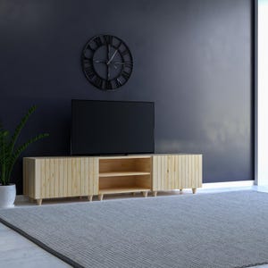 May include: A light wood TV stand with vertical slat detailing, featuring open shelving and a flat-screen TV. A large black clock hangs on the dark blue wall above the stand. A gray rug and a potted plant complete the room's decor.
