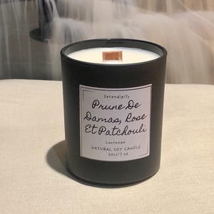 Op de afbeelding: Een zwarte cilindrische kaars met een houten lont. Het witte etiket draagt de tekst "Serendipity Prune De Damas, Rose Et Patchouli NATURAL SOY CANDLE 20cl/7 oz". De kaars staat op een neutrale ondergrond.