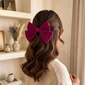 以下が含まれることがあります： 大きなマゼンタ色のリボンヘアクリップが、ある人の茶色の髪を飾っています。リボンは、ベージュのセーターと額入りの写真や装飾品が置かれた棚を背景にしています。全体的な雰囲気は柔らかく、女性的です。