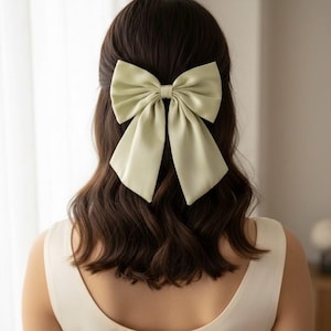 May include: A large, pale green satin bow hair clip. The bow is centered on dark brown wavy hair. The person is wearing a cream-coloured dress. The bow is a statement piece, adding a touch of elegance to the hairstyle.