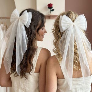 Könnte beinhalten: Zwei Frauen mit weißen Haarschmuck-Schleifen aus Tüll. Die Schleifen sind groß und luftig, mit langen, fließenden Schleiern. Sie tragen passende beige Tanktops. Im Hintergrund ist eine rote Rose zu sehen.