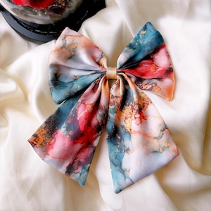May include: A fabric hair bow featuring an abstract watercolour pattern in shades of teal, deep red, and gold. The bow has a smooth, satin-like texture with a central knot detail, showcasing a vibrant, multi-coloured marbled design throughout.