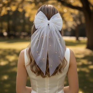 Peut inclure: Un voile blanc en forme de nœud en tulle orné de perles dorées. Le voile est fixé à l'arrière de la tête et retombe sur les cheveux. L'arrière-plan est une scène extérieure floue avec des arbres.