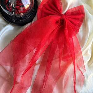 May include: A large, bright red tulle bow with long, flowing streamers, next to a preserved red rose in a glass dome. The bow is the main feature, with a full, layered design. The rose sits on a black base with a black ribbon that says "Just for you".