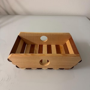 May include: A rectangular wooden tray with a striped interior and rounded cut-out handles. The tray is constructed from light and dark brown wood, with a scalloped edge detail. The design is simple and rustic.