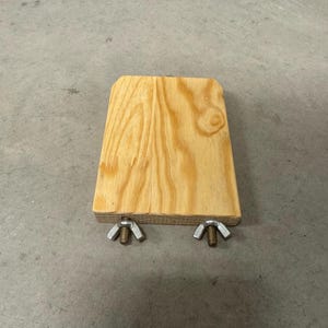 May include: A rectangular wooden block with rounded top corners, attached to two metal wing nuts. The wood grain is visible. The object is on a gray concrete surface.