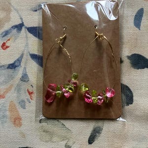 May include: A pair of gold-colored wire hoop earrings. Each earring is adorned with a cluster of pink and green gemstone beads. The earrings are displayed on a brown cardboard backing, sealed in a clear plastic bag.