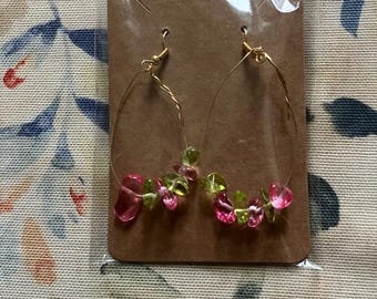 May include: A pair of gold-coloured wire hoop earrings. Each earring features a cluster of pink and green gemstone beads. The earrings are presented on a brown cardboard backing, sealed in a clear plastic bag.
