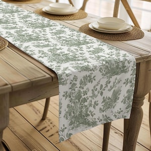 May include: A white and green floral patterned table runner is displayed on a wooden dining table. The runner features a detailed botanical design. The table is set with plates, bowls, and woven placemats, creating a rustic and elegant dining scene.