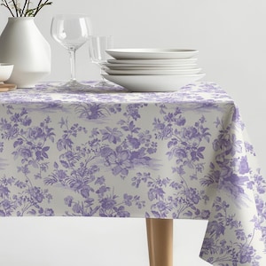 May include: A white tablecloth with a purple floral pattern. The tablecloth is laid on a table with a vase of white flowers, a bowl, and a stack of white plates.