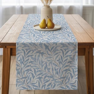 Peut inclure: Chemin de table à motif de feuilles bleu clair et blanc sur une table en bois. Le motif répété représente des feuilles stylisées. Un vase avec des plantes séchées et une assiette avec deux poires sont sur la table.
