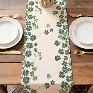 May include: A cream-colored table runner with a repeating pattern of green shamrocks. The runner is centered on a wooden table, with white plates, gold cutlery, and a woven basket visible.