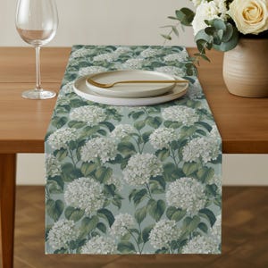 May include: A rectangular table runner with a floral design of white hydrangea flowers and green foliage on a muted teal backdrop. The runner is displayed on a wooden table, with a wine glass, plates, and a vase of flowers.